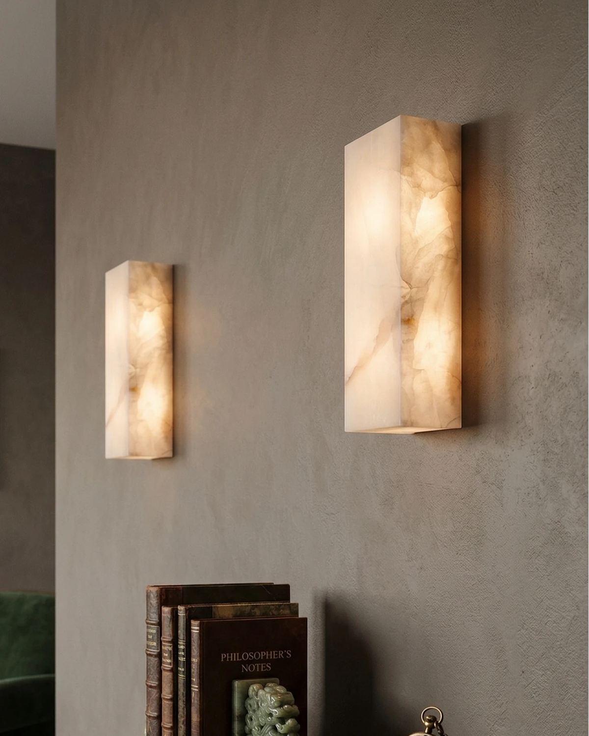 Two alabaster wall sconces ALABA No.03 lit on a plaster wall corner with a stack of books on the floor