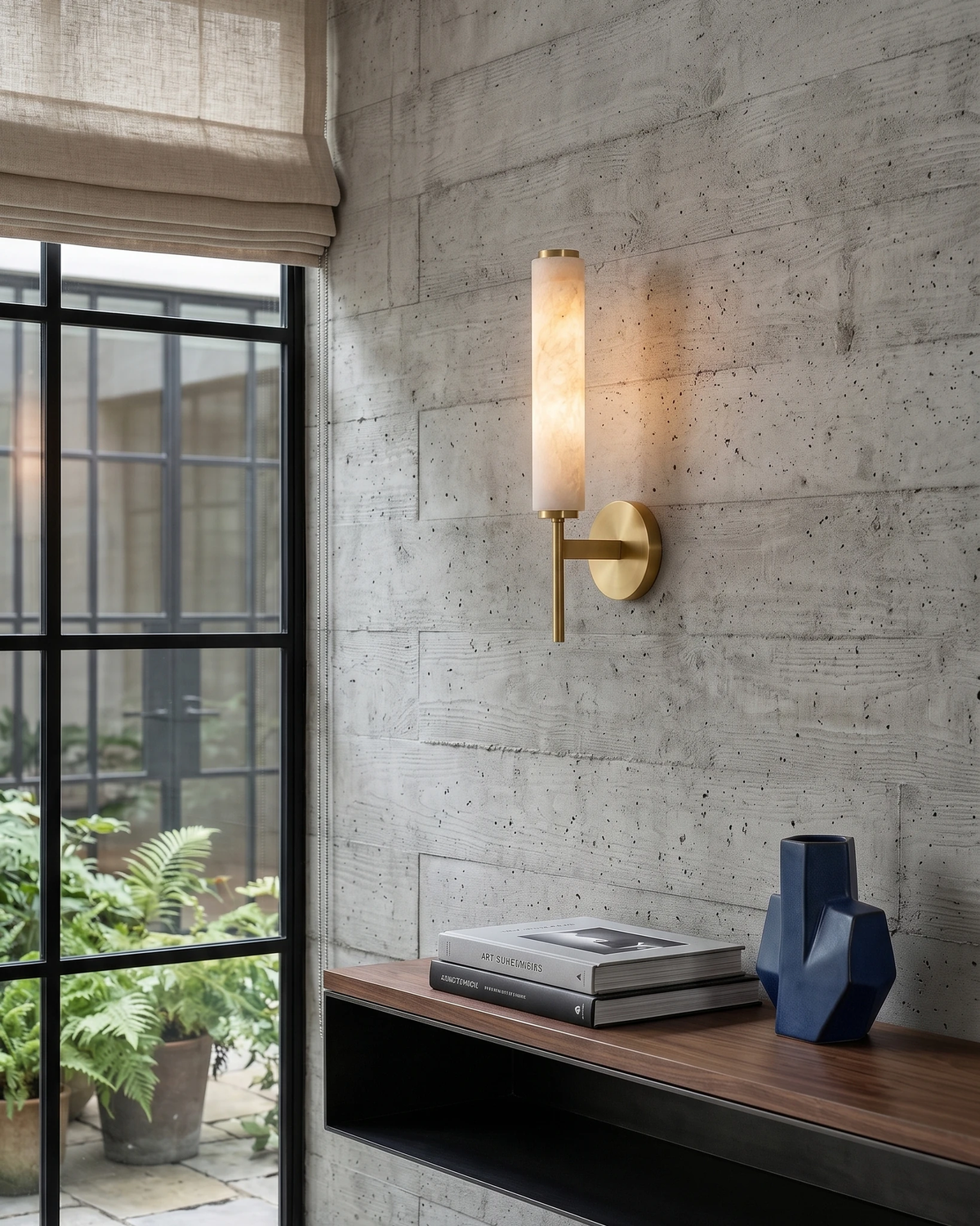 Alabaster wall sconce ALABA No.01 with brass bracket, lit on a concrete wall beside a steel-framed window