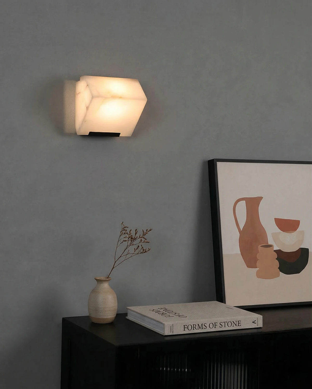 Alabaster wall sconce ALABA No.05 solo, single faceted block lit on a grey wall above a black console with a print
