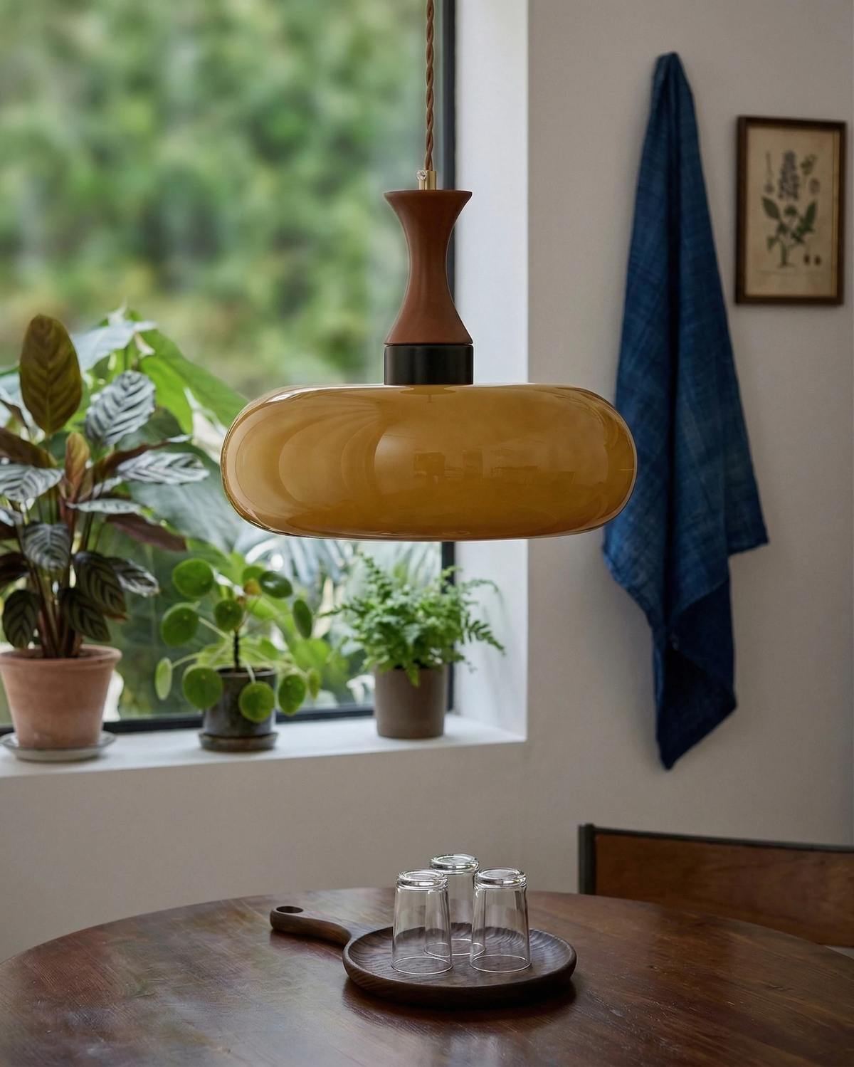 Amber glass pendant light OVIA No.04, disc shade over a round wood table by a window full of plants