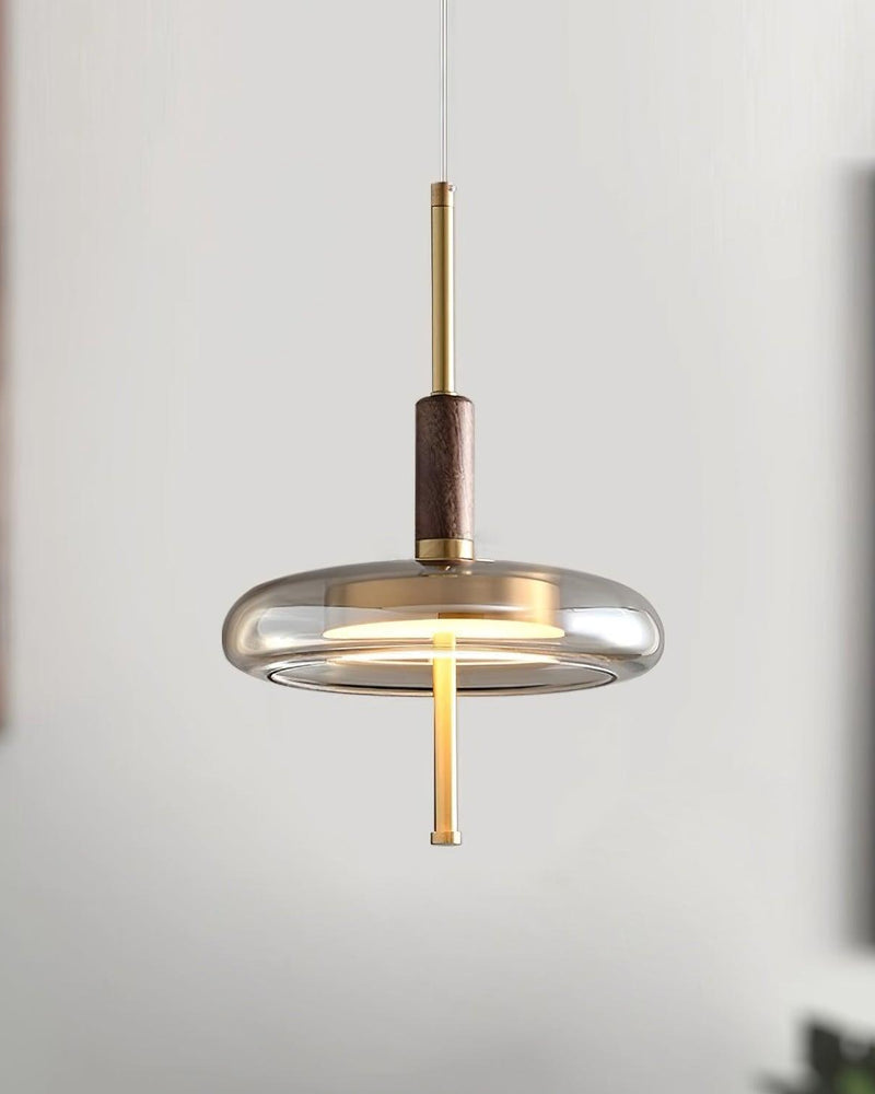Amber glass pendant light NOCE No.01 lit, walnut and brass stem above a smoked glass saucer, blurred gray backdrop