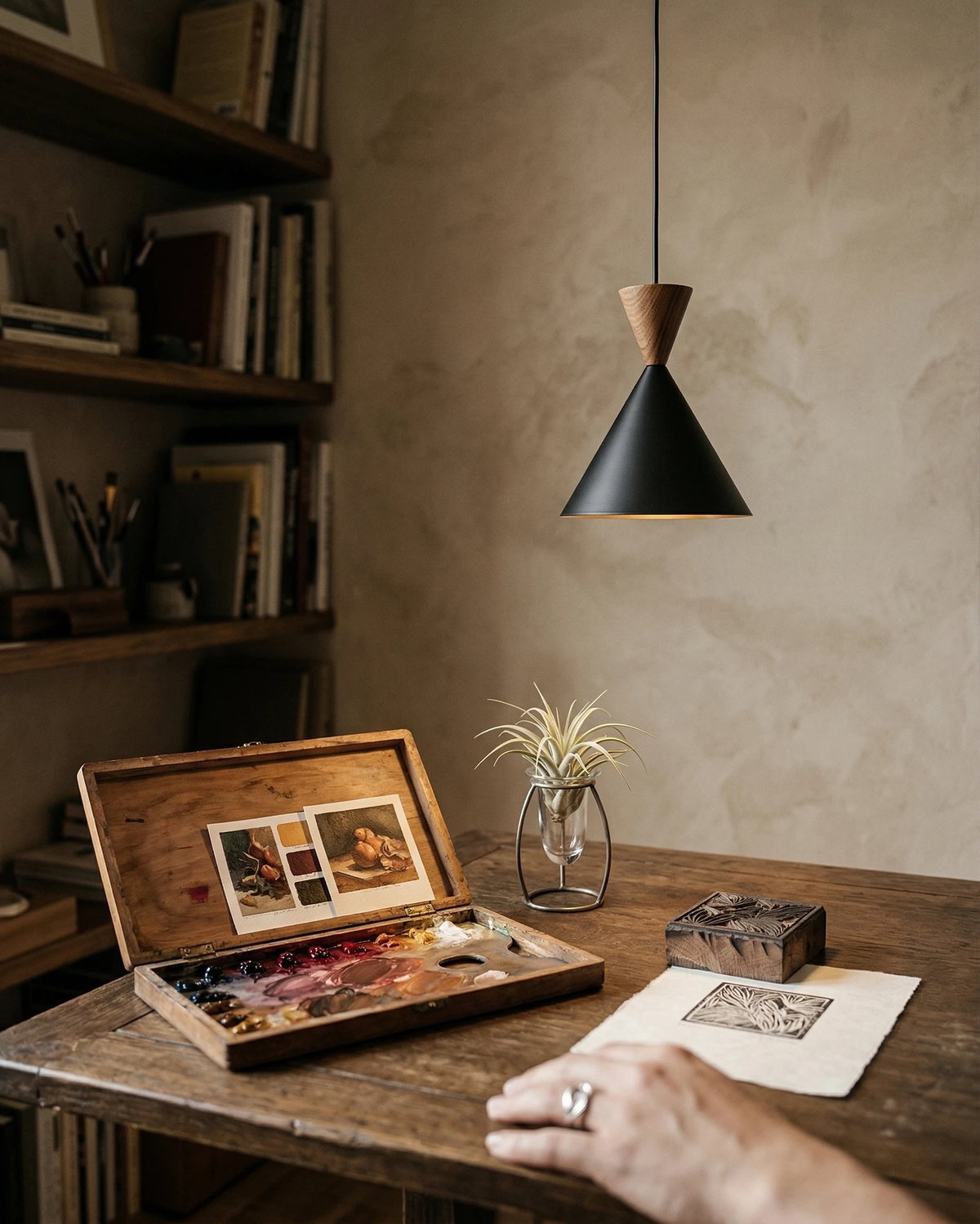 Brass pendant light TARO No.01 in matte black hanging above a wooden desk with a paint box and a hand at work