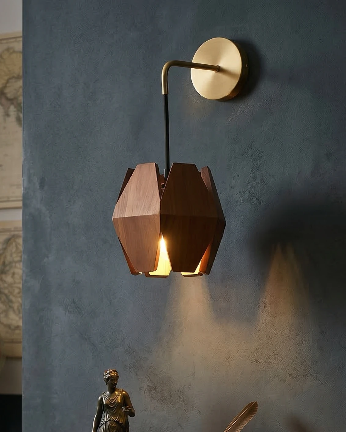 Brass wall sconce NODO No.01 with a faceted walnut shade glowing on a dark plaster wall beside a bronze figurine