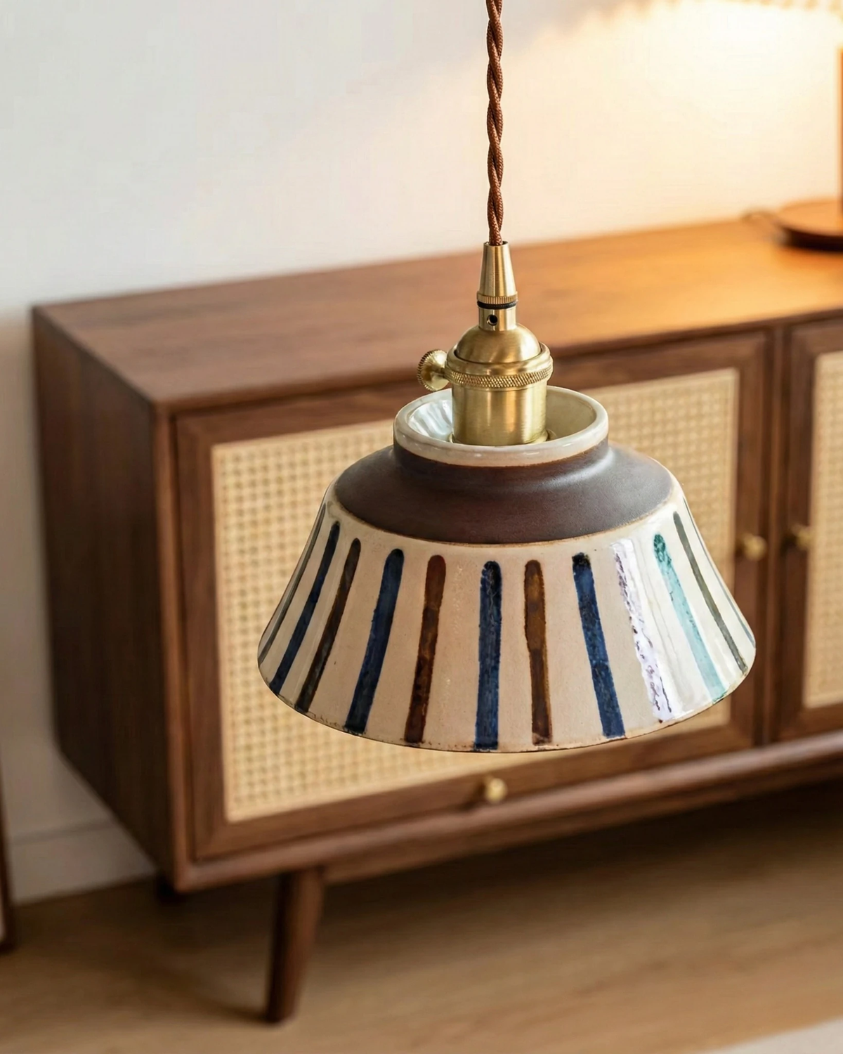 Ceramic pendant light MELI No.05 in ribbed earth stripes lit in front of a walnut and cane sideboard