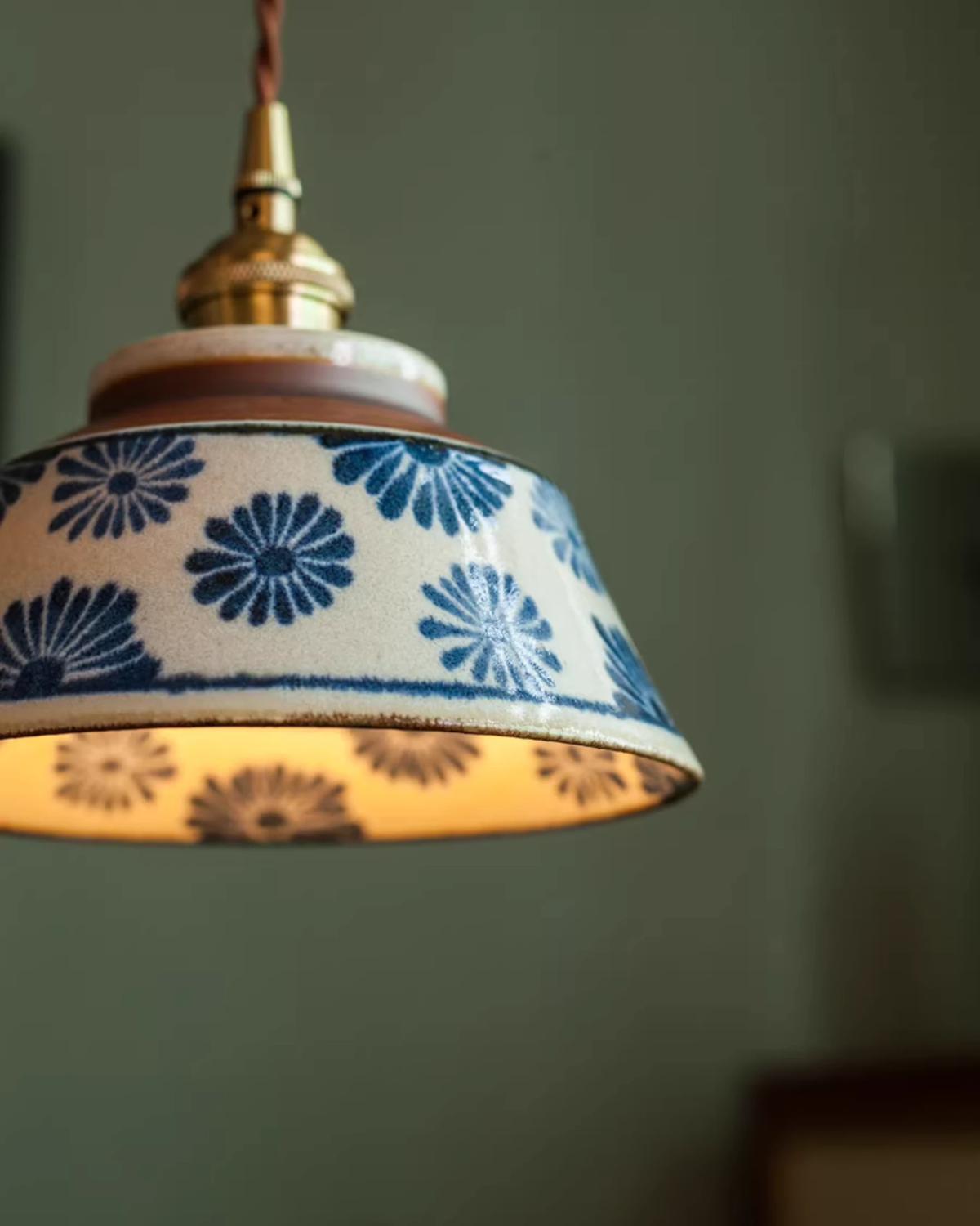 Close-up of the indigo floral bell and brass fitting of the MELI No.05 ceramic pendant light, lit against a green wall