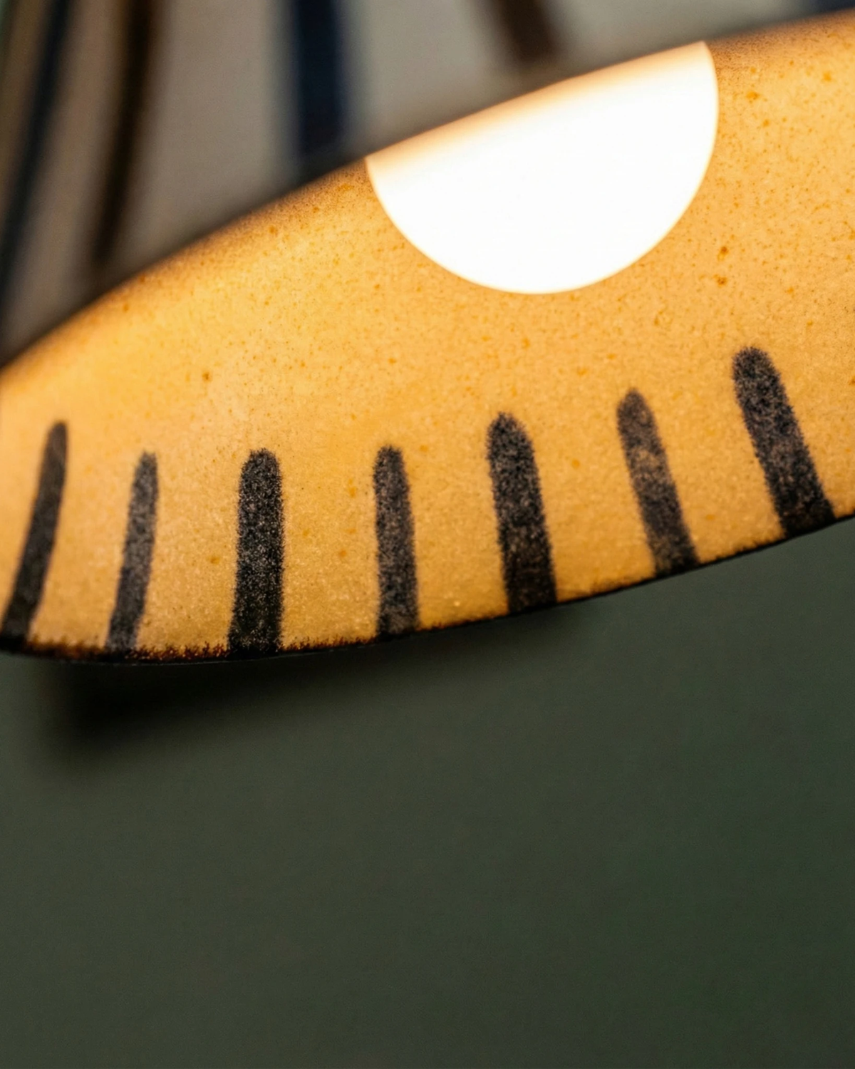 Close-up from below of the MELI No.05 ceramic pendant light showing the bulb glowing inside the striped bell