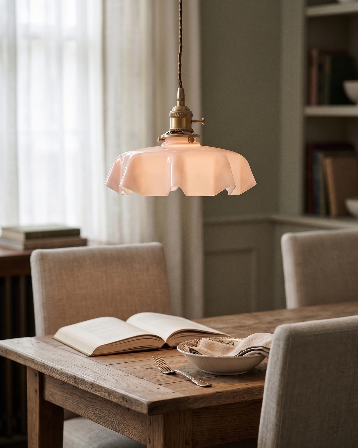 Ceramic pendant light CORA No.01 in dusty rose switched on above a wooden dining table with an open book
