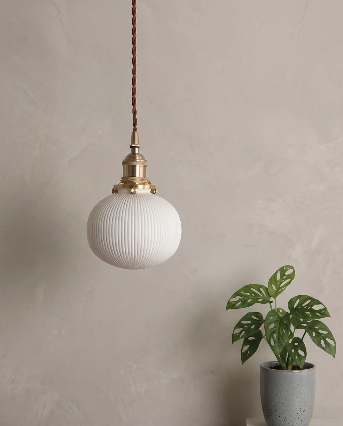Ceramic pendant light SERA No.01 pebble shade hanging on a plaster wall beside a potted monstera plant