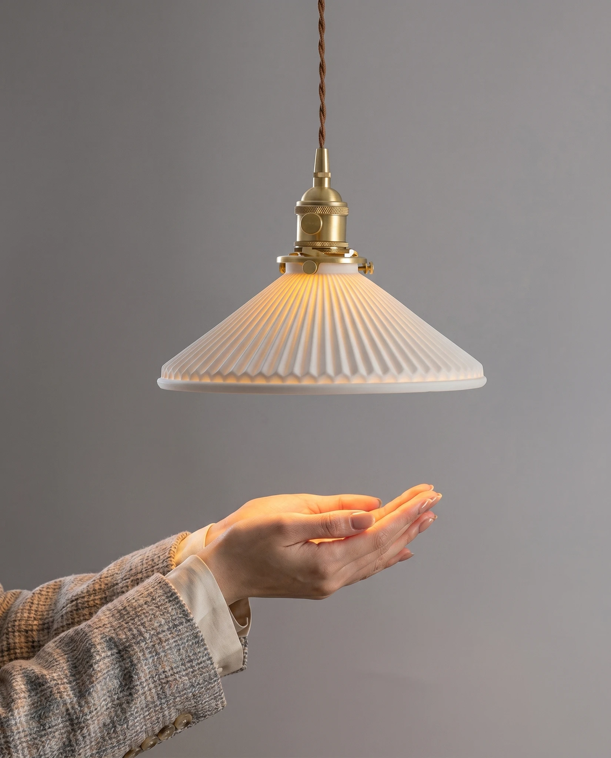 Ceramic pendant light CORA No.02 lit against a gray wall with a hand cupped beneath the pleated shade