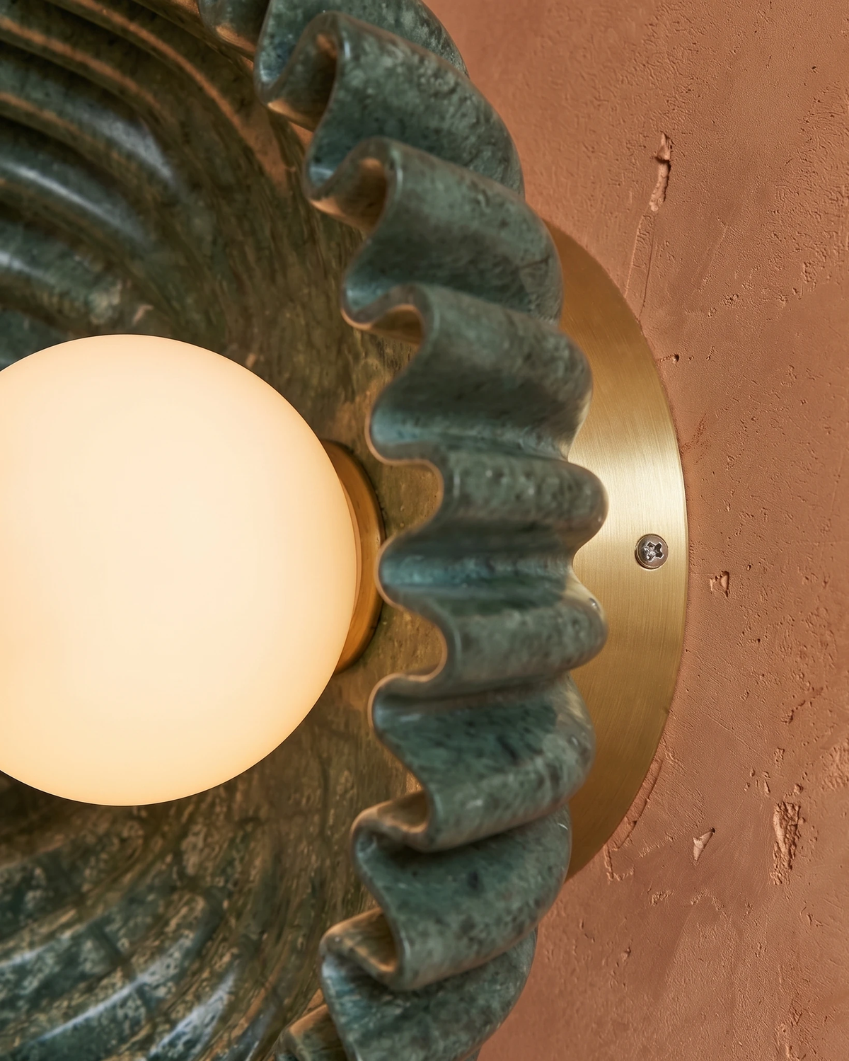 Close-up of the lit opal globe and green scalloped ceramic rays of the RAGGI No.01 ceramic wall sconce on terracotta plaster