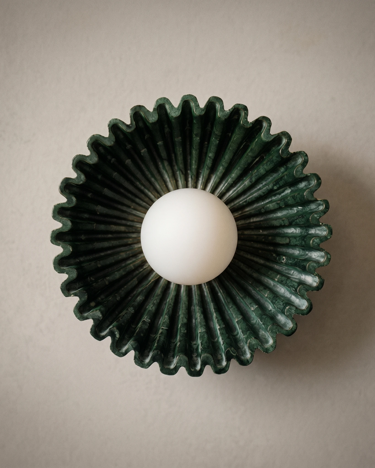 Ceramic wall sconce RAGGI No.01 in green, front view of the sunburst ring and opal globe on a beige plaster wall, unlit