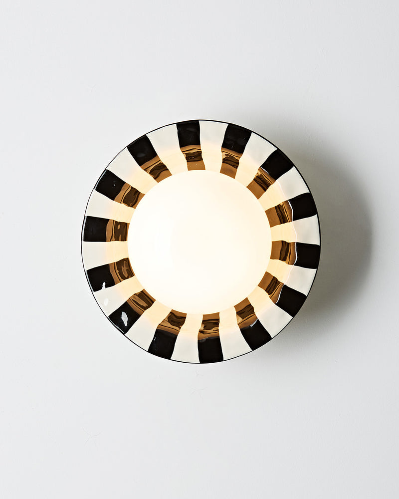 Ceramic wall sconce OLEA No.01 viewed head-on, lit opal globe ringed by a black and white striped ceramic corolla