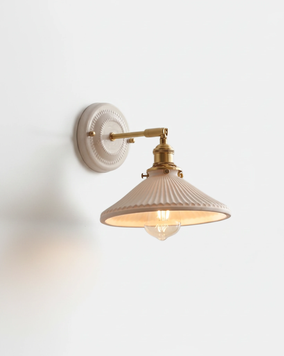 Ceramic wall sconce RIGA No.04, white ribbed coolie shade on an antique brass swing arm with the bulb lit