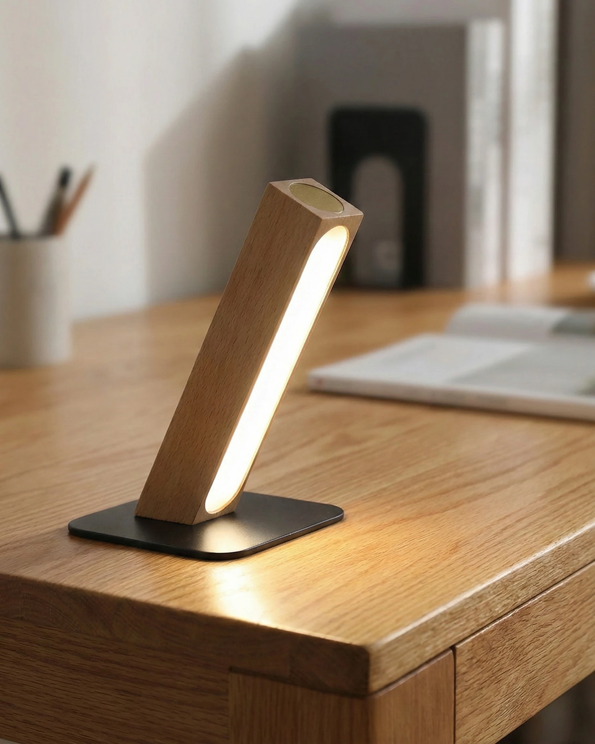 Cordless desk lamp RAMO No.01 in light beech, lit on a wooden desk with a pencil cup