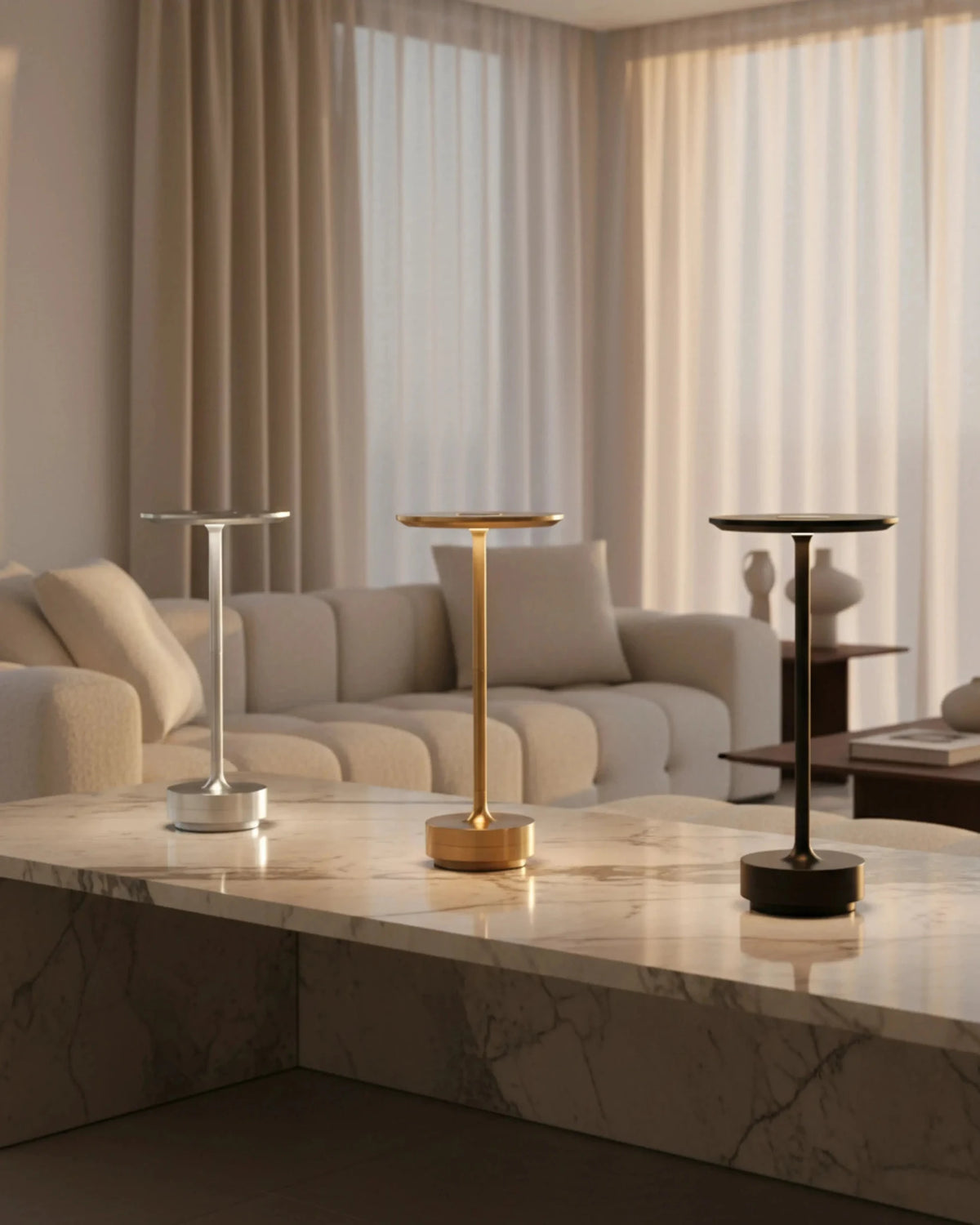 Three cordless desk lamps LUMA No.05 in silver, brass and black lit on a stone coffee table by a cream sofa