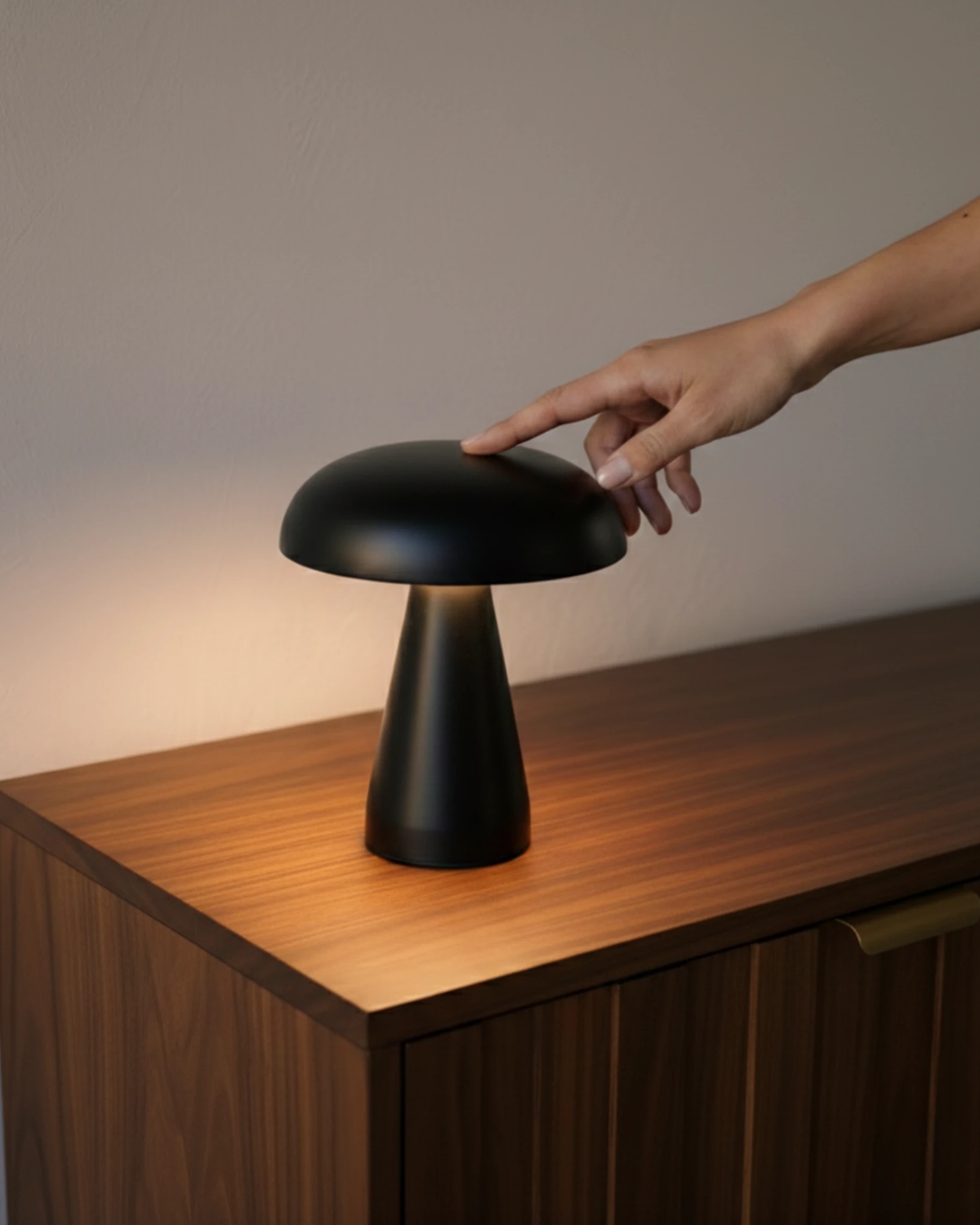 Hand touching the cordless mushroom lamp LUMA No.04 in black, lit on the corner of a walnut cabinet