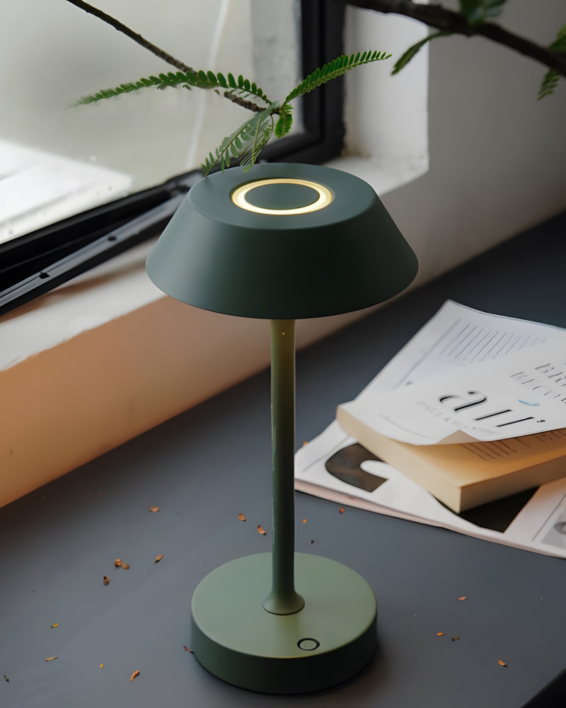 Cordless mushroom lamp CIMA No.01 in forest green lit on a dark desk by a window with magazines and a fern