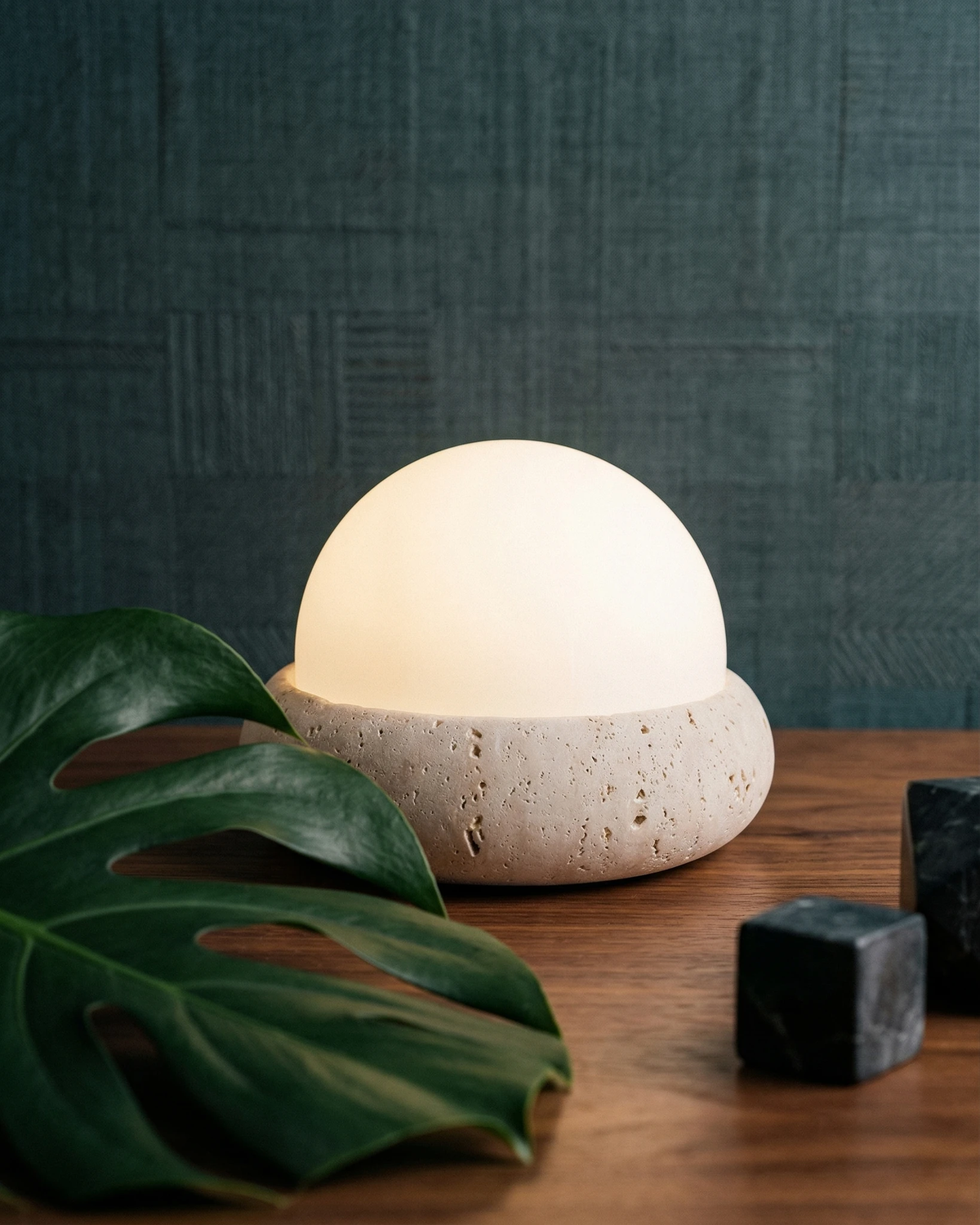Cordless outdoor table lamp LUCE No.01 glowing on a walnut table next to a monstera leaf against a dark blue wall