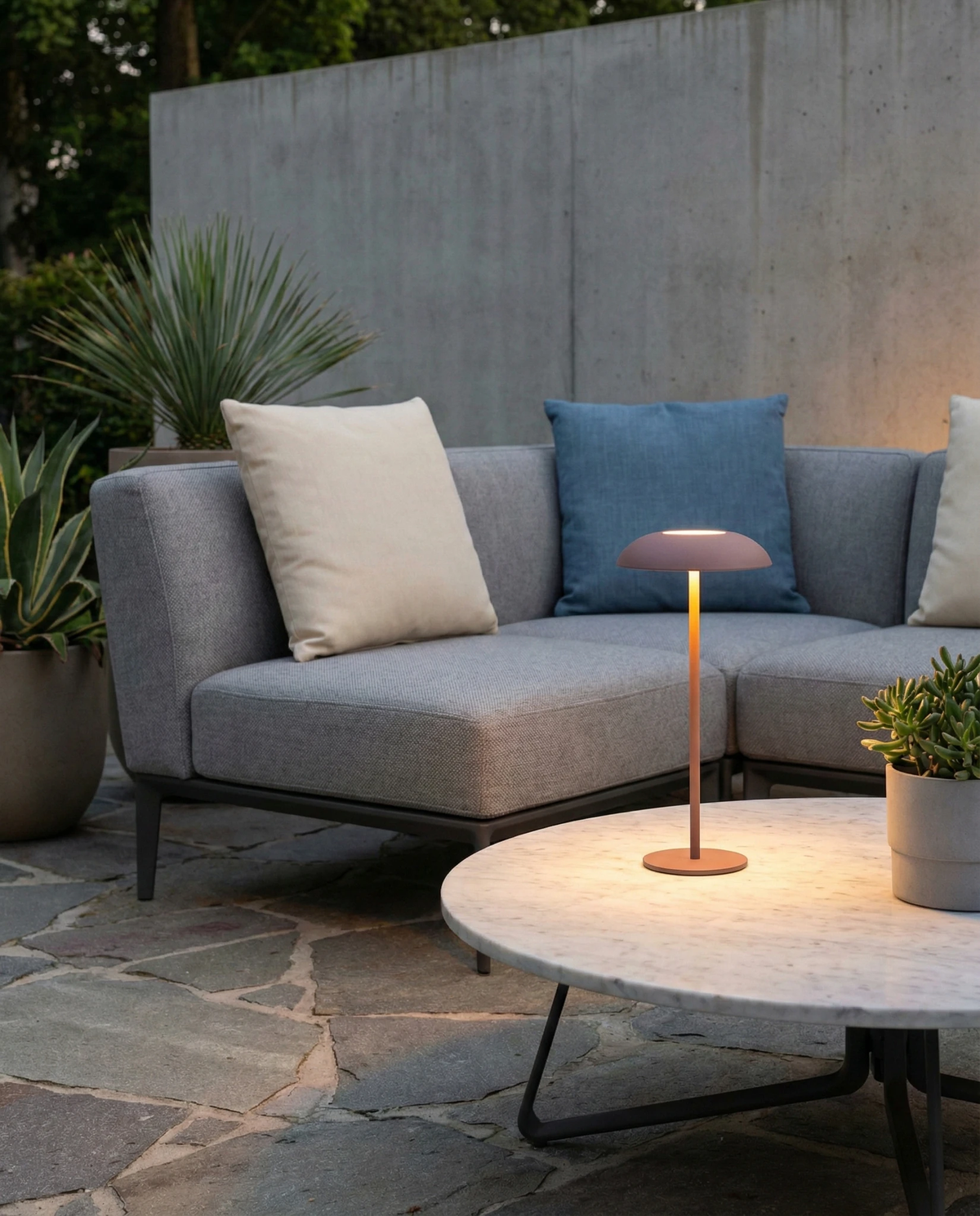 Cordless table lamp PESCARA No.01 in brown lit on an outdoor coffee table in front of a gray sectional sofa