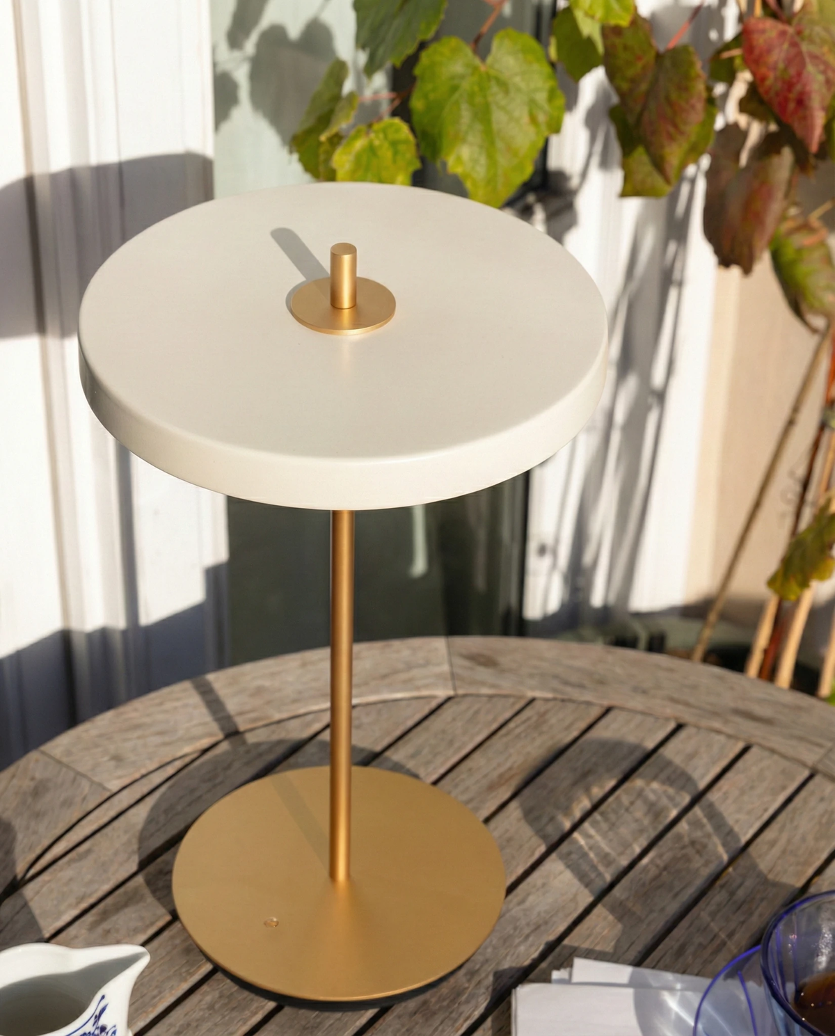 Cordless table lamp SALA No.01 in matte white on a wooden patio table in sunlight beside potted plants
