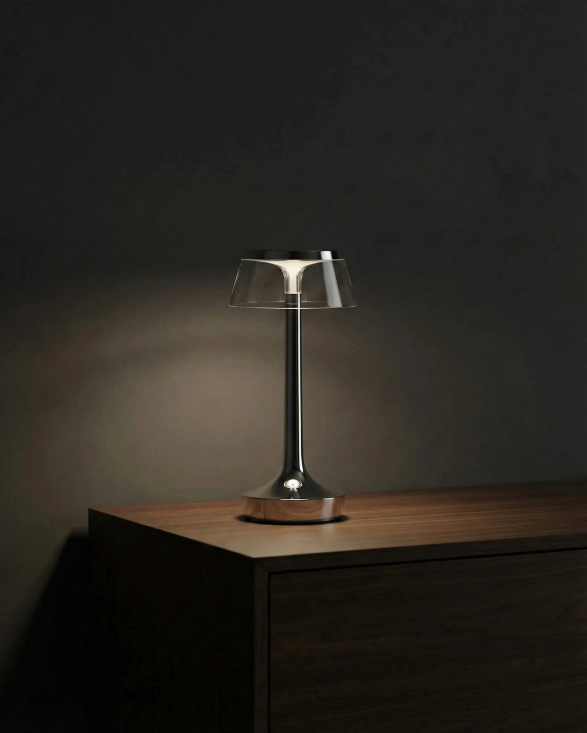 Cordless table lamp ZARA No.01 in chrome switched on atop a dark wooden cabinet in a dim room