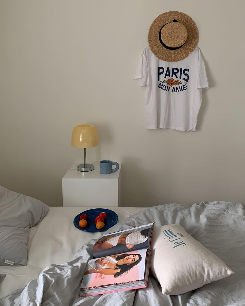 Glass table lamp VENEZIA No.01 on a white nightstand beside a bed, with a t-shirt and straw hat on the wall