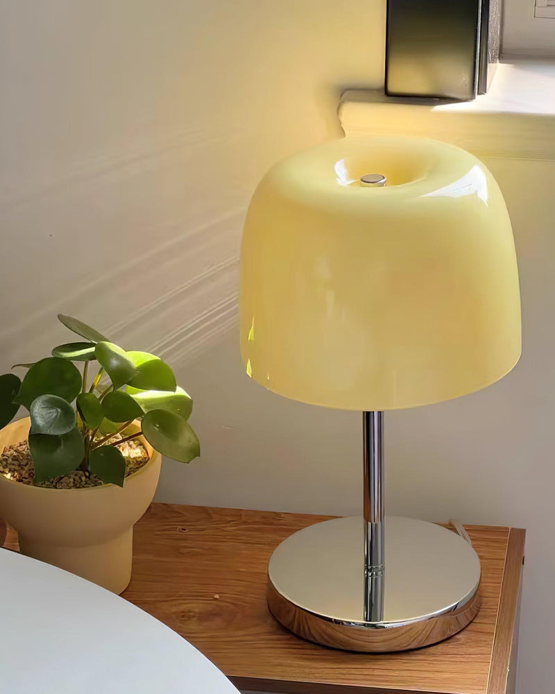 Glass table lamp VENEZIA No.01 switched on, glowing pale yellow on a wooden table beside a potted plant
