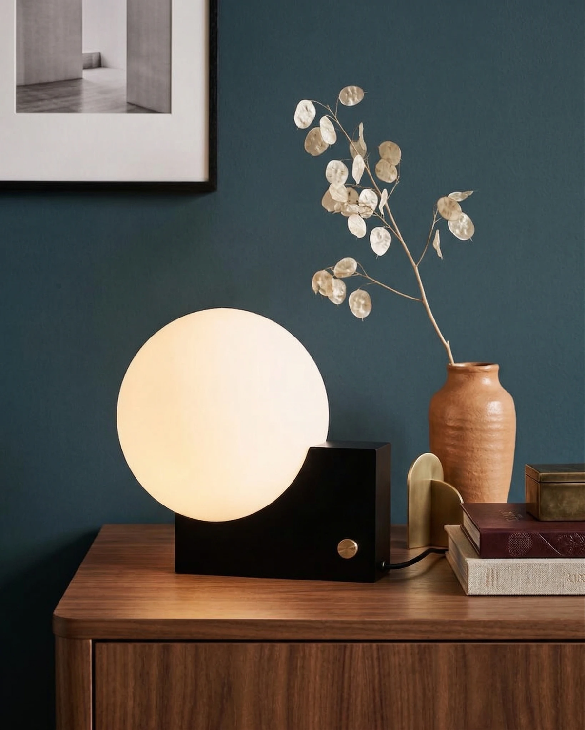 Globe desk lamp NOVA No.01 in matte black lit on a walnut sideboard with a vase against a dark green wall