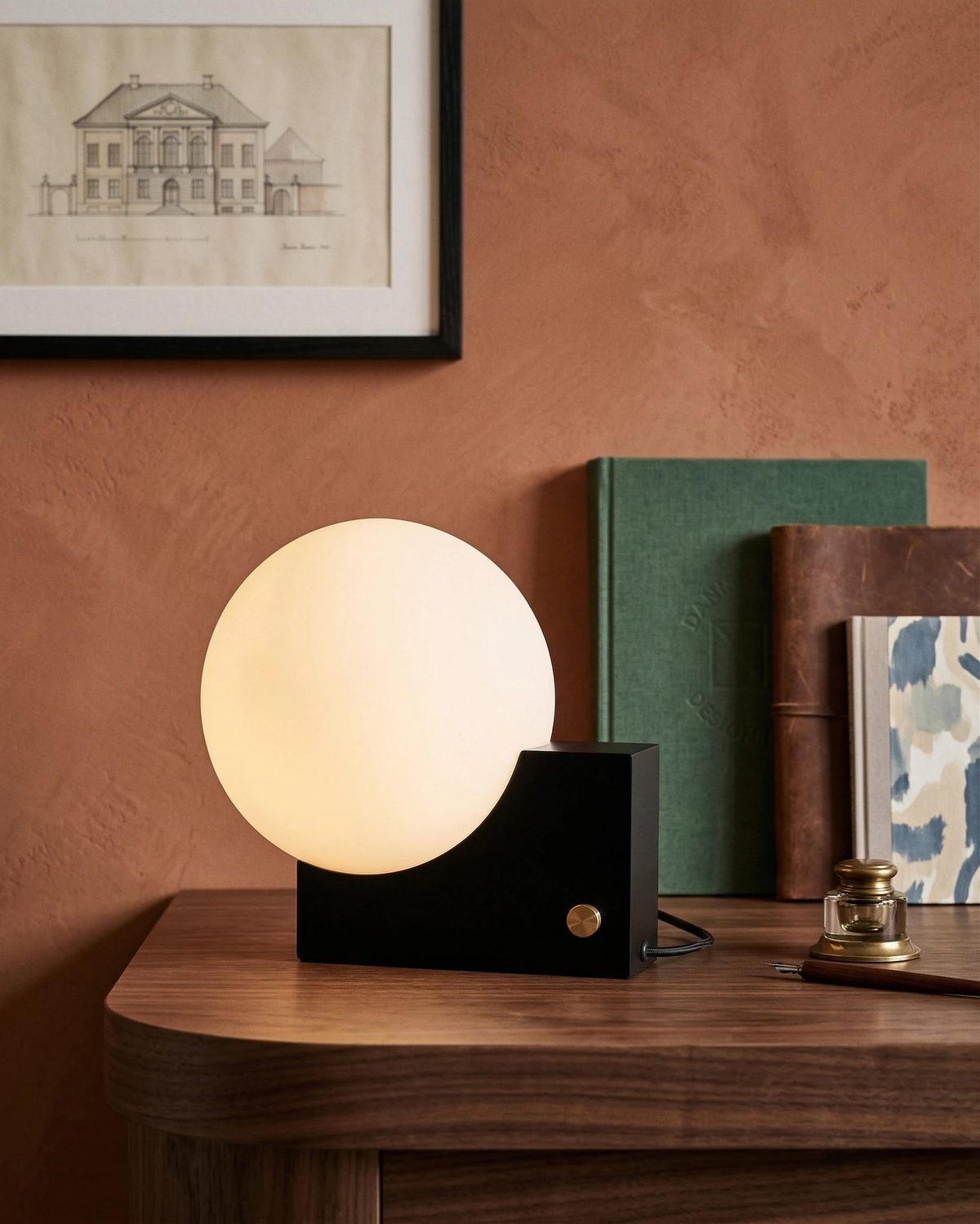 Globe desk lamp NOVA No.01 in matte black lit on a walnut desk with books against a terracotta wall