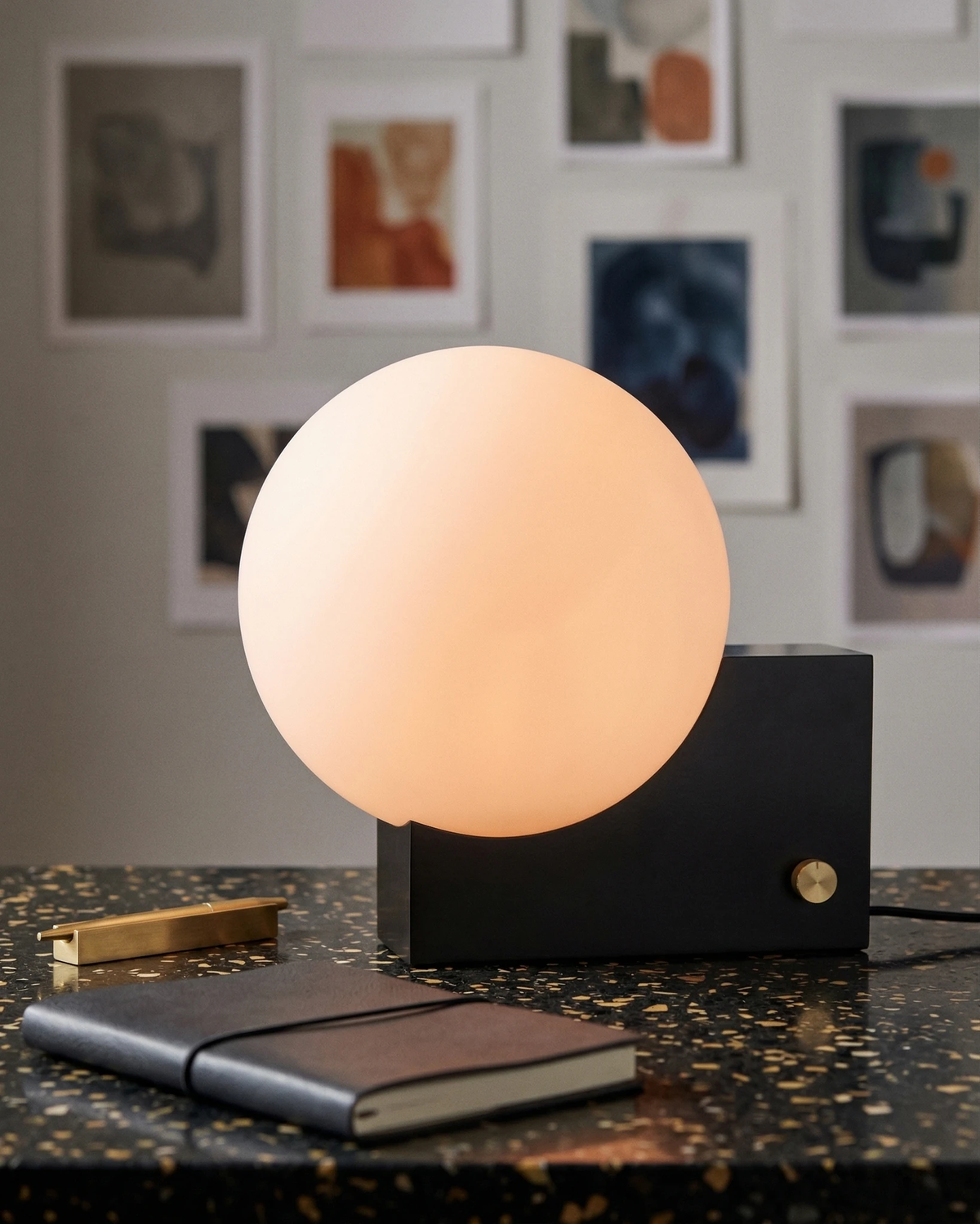 Globe desk lamp NOVA No.01 in matte black lit on a terrazzo desk with a notebook beneath a gallery wall