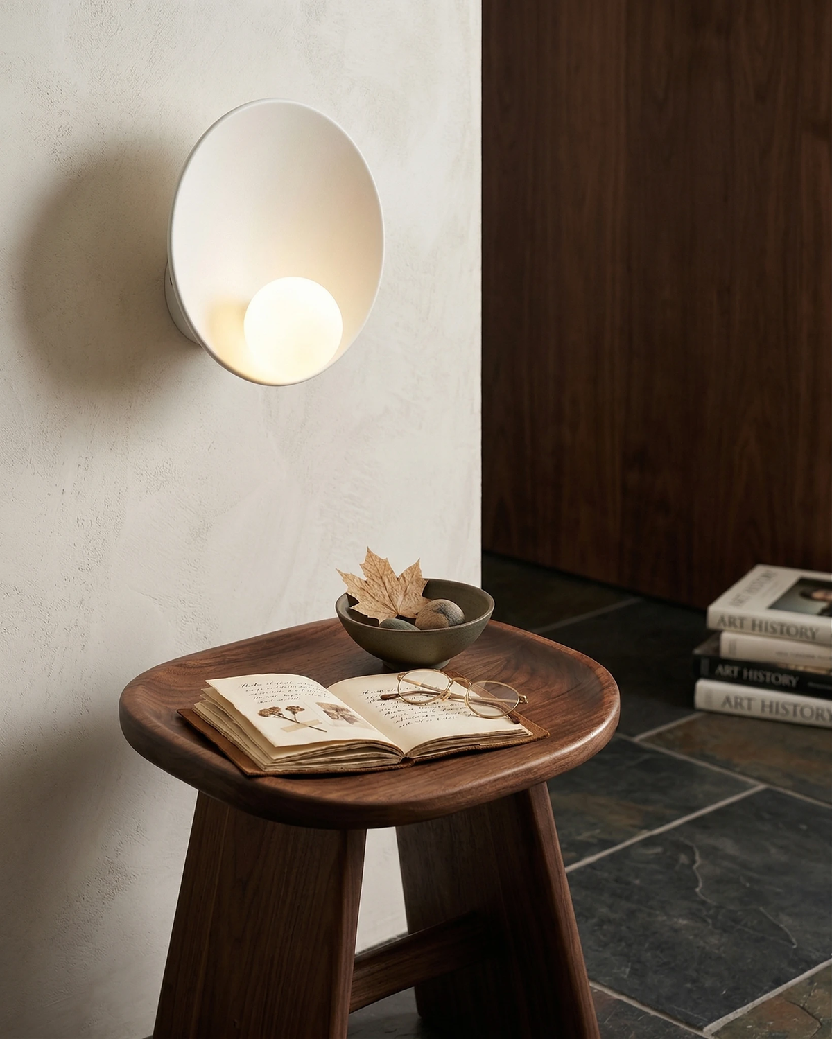 Globe wall sconce OASI No.01 in pure white, lit on a plaster wall above a wooden stool with an open book