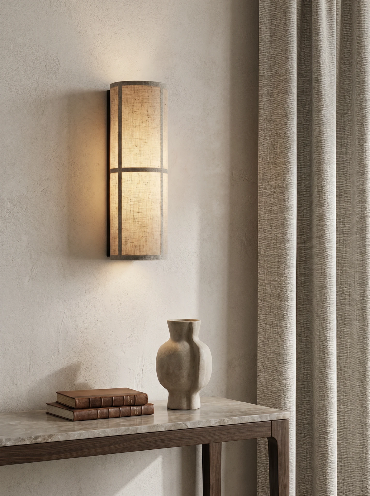 Linen shade wall sconce FUMO No.01 lit above a stone console with a vase and books beside a curtain