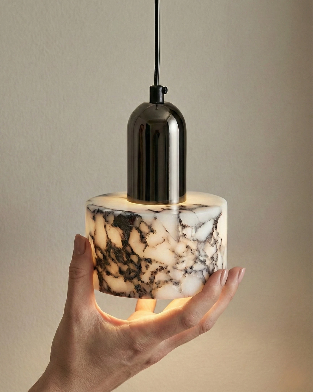 Hand cupping the lit marble cylinder of the MARMI No.02 marble pendant light below its black chrome socket