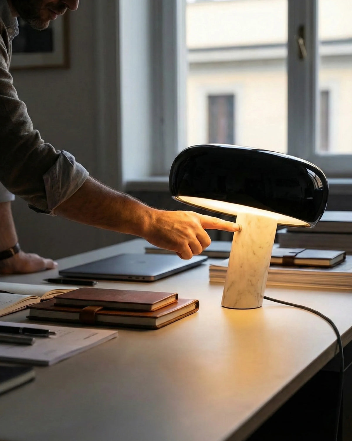 Marble table lamp BRUNO No.01 with black enamel shade, switched on on a desk as a man reaches for a notebook by the window