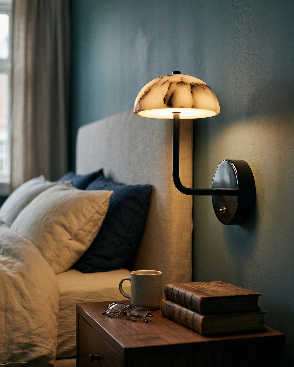 Marble wall sconce NUVOLA No.01 lit beside a bed with blue pillows and a nightstand with books and a mug