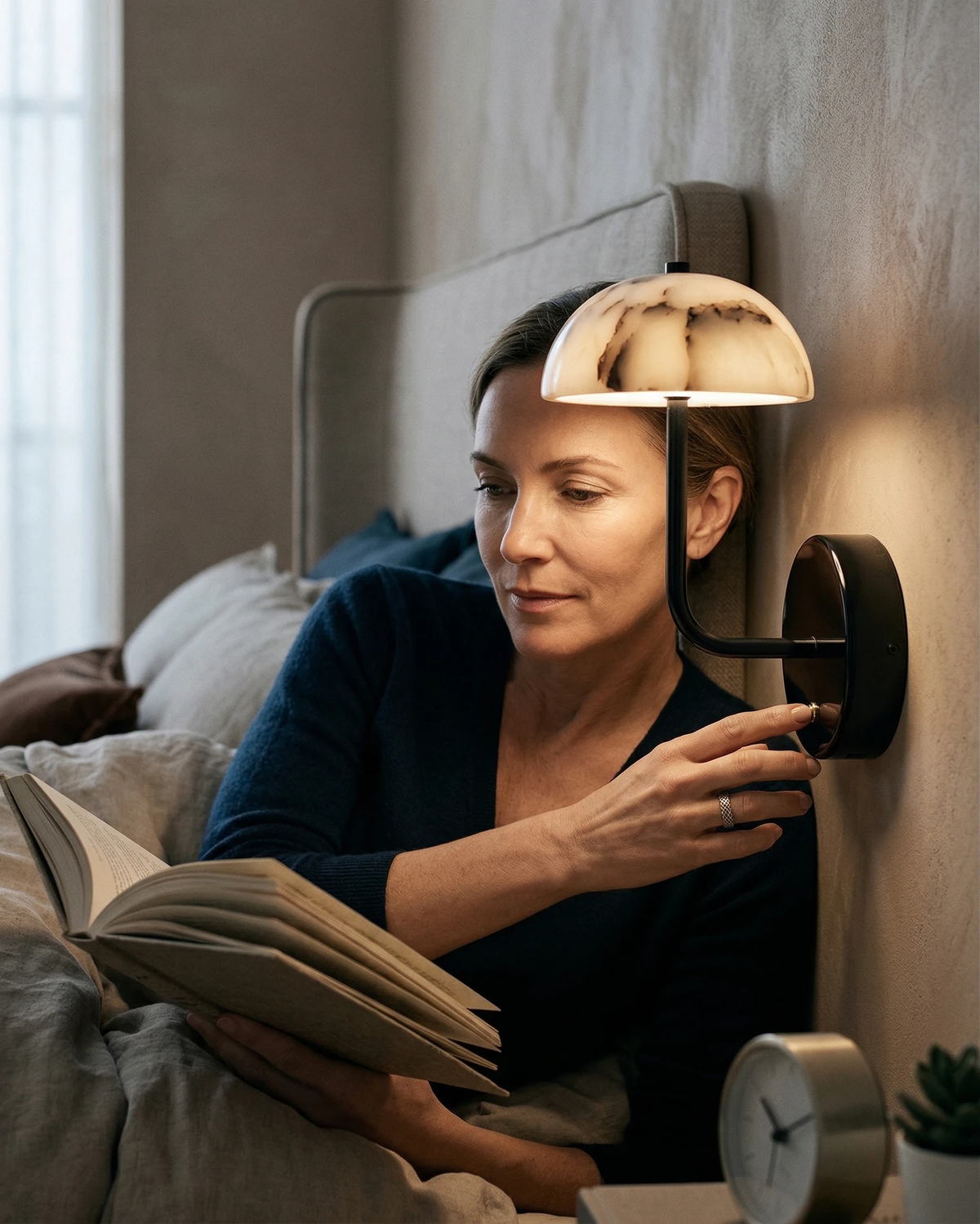 Marble wall sconce NUVOLA No.01 lit above a woman reading in bed and reaching for the switch