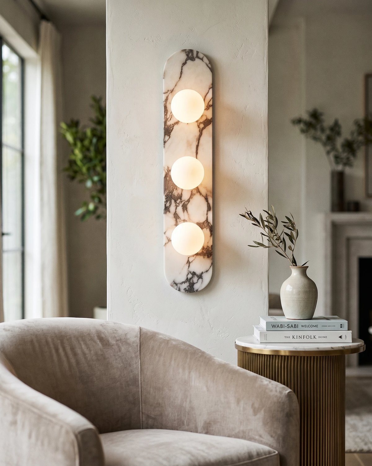 Triple globe marble wall sconce AVOLA No.01 lit on a white wall behind a beige armchair and side table of books