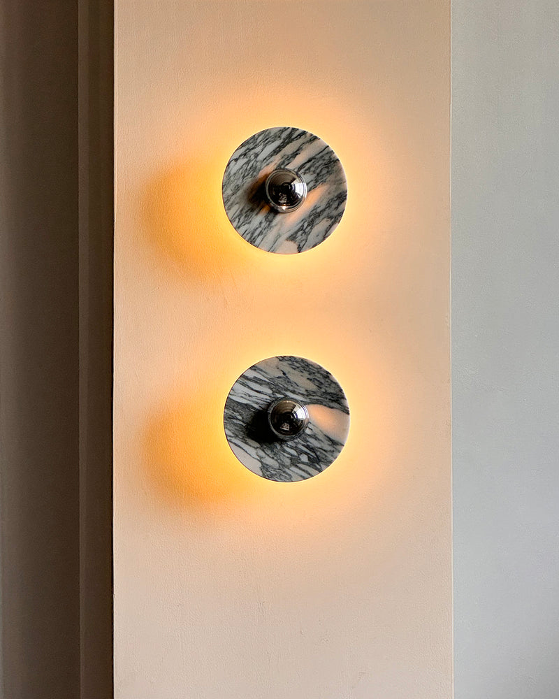 Marble wall sconce LATTE No.01, two chrome units stacked vertically and switched on with a warm halo on a beige wall