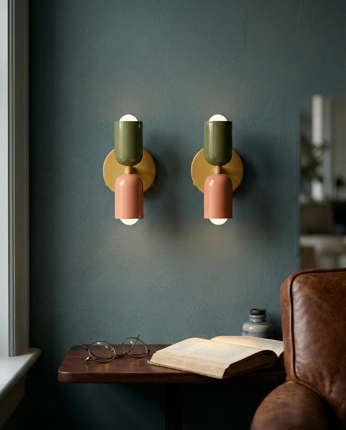 Two modern wall sconces MURO No.02 in sage and pink, switched on against a teal wall above a small reading shelf