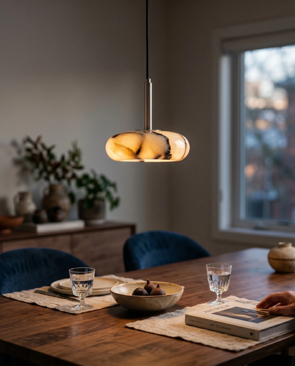 Onyx pendant light TIBER No.01 switched on above a walnut dining table with blue velvet chairs at dusk
