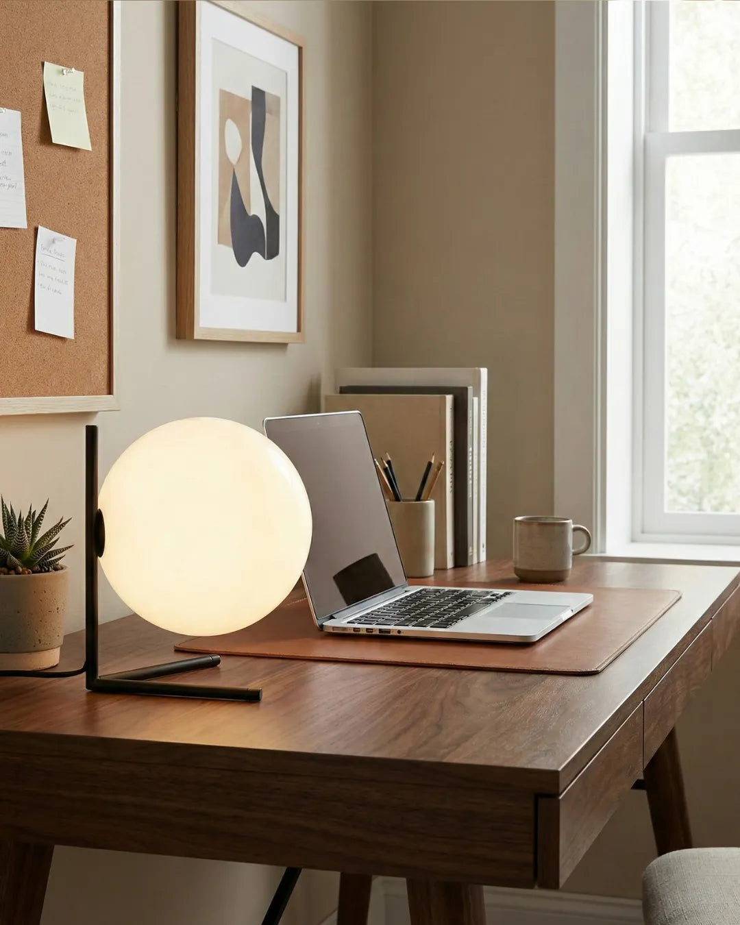Opal globe table lamp ARIA No.01 lit on a walnut desk beside a laptop under a pinboard by a window