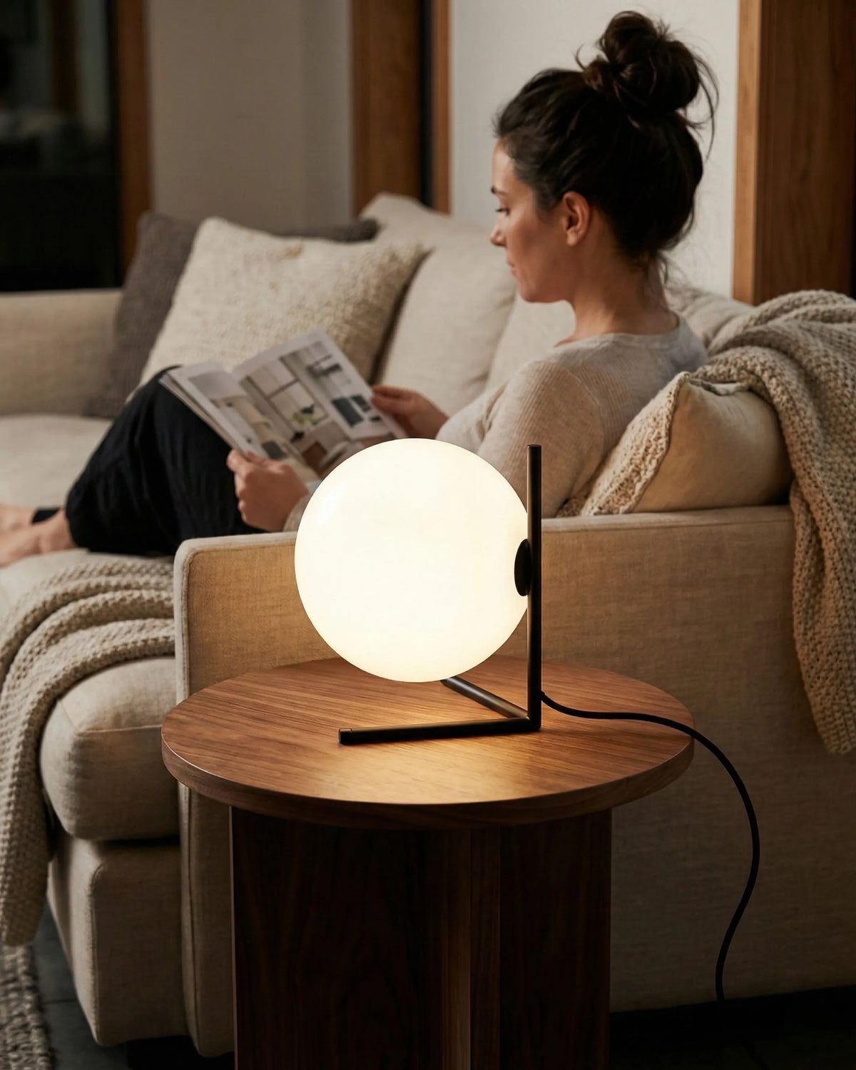 Opal globe table lamp ARIA No.01 lit on a round side table beside a woman reading on a cream sofa