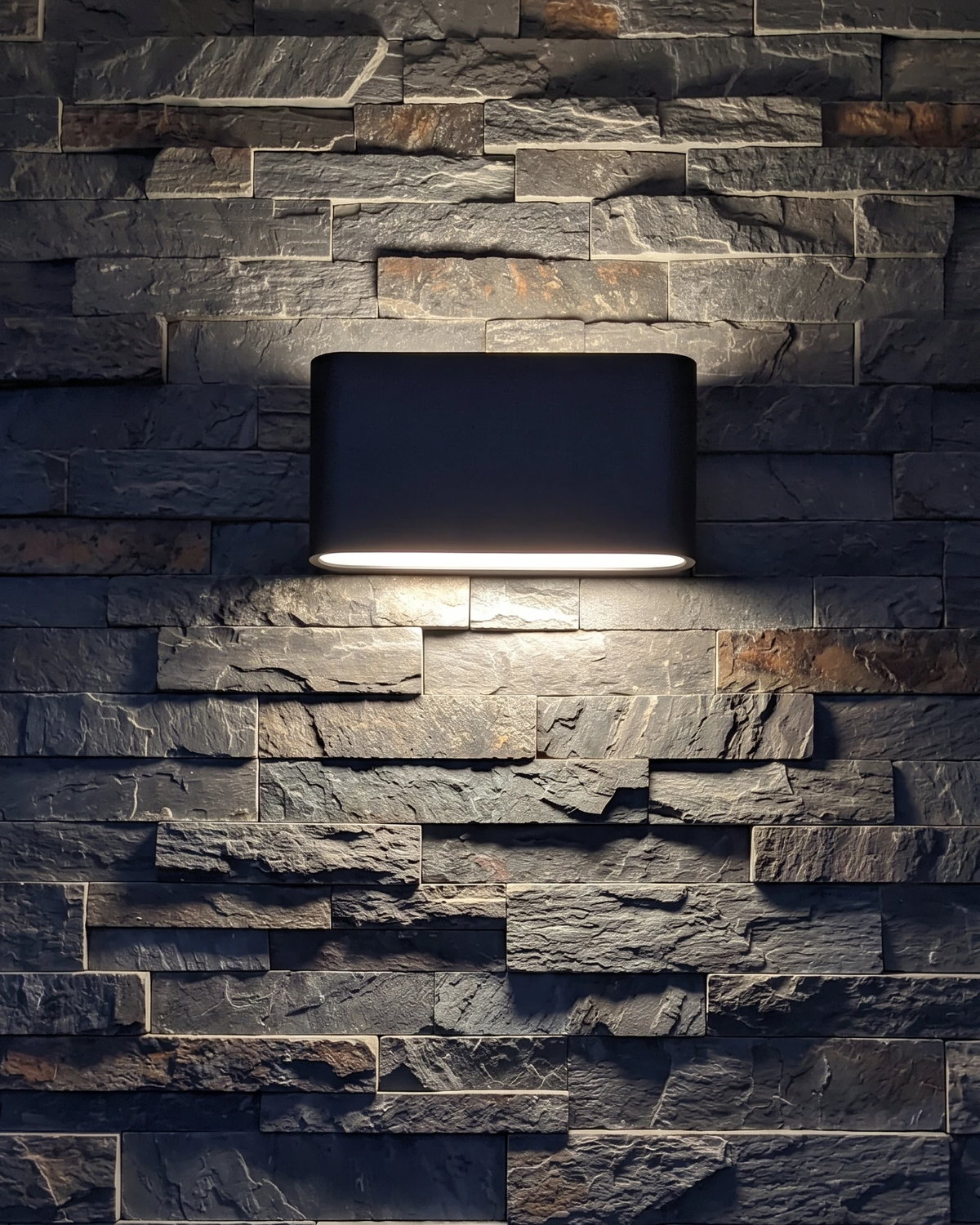 Outdoor wall sconce PISA No.01 in deep black switched on against a dark stacked slate wall at night