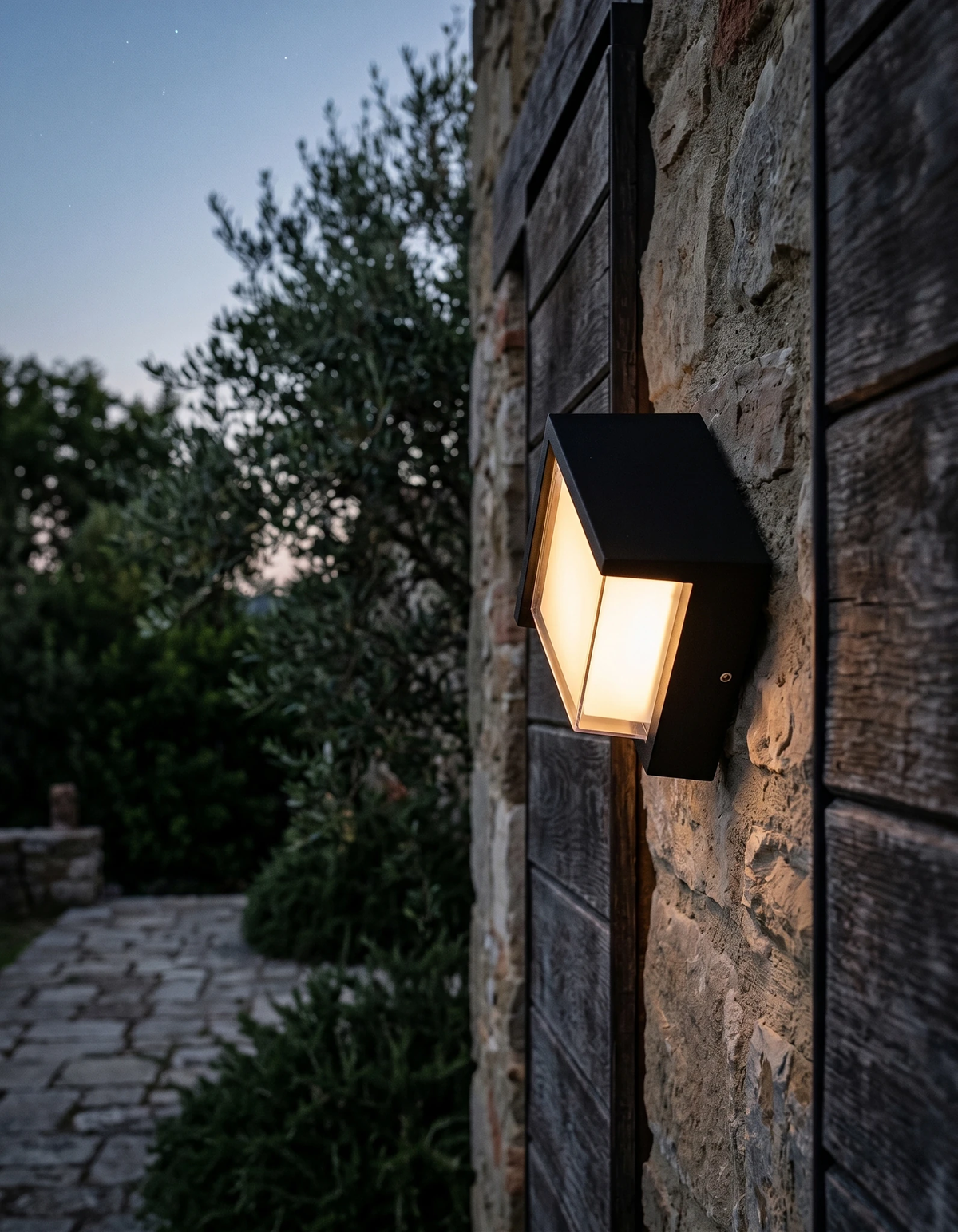 Outdoor wall sconce FARA No.02 lit on a rough stone wall at dusk above a cobbled path and garden