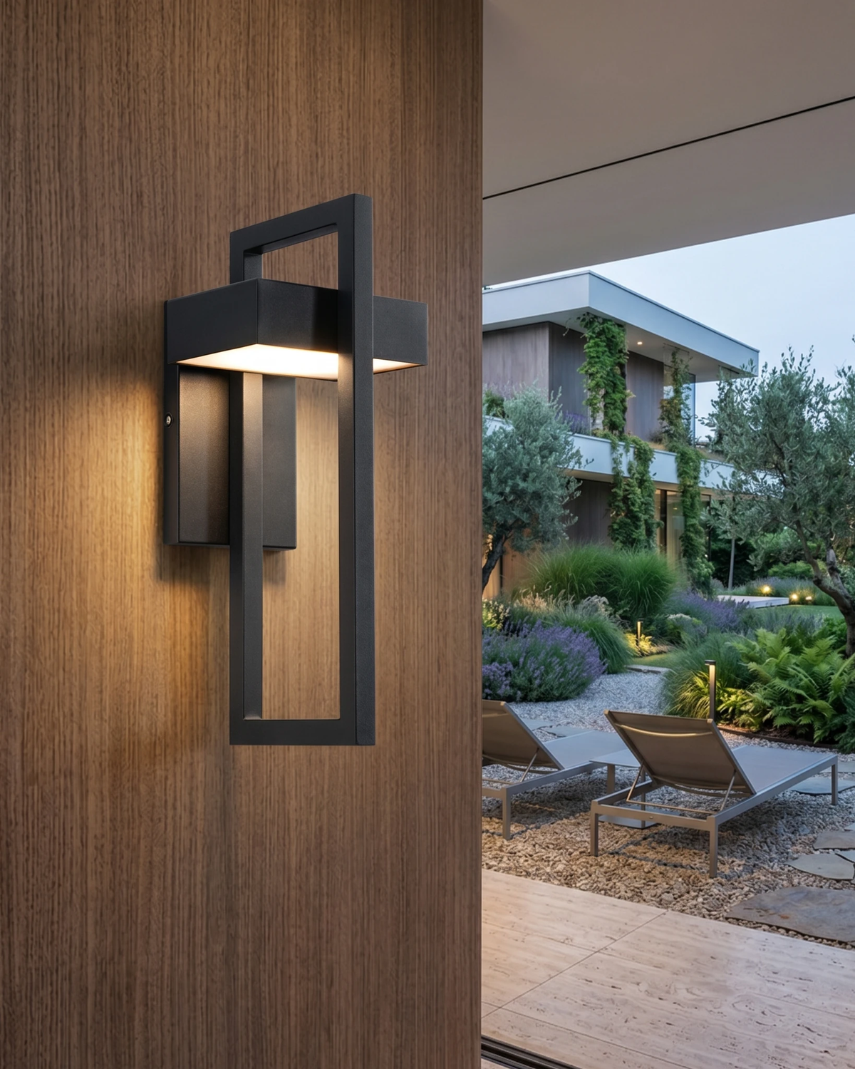 Outdoor wall sconce FERRO No.01 lit on a wood-clad wall beside an opening to a garden with sun loungers