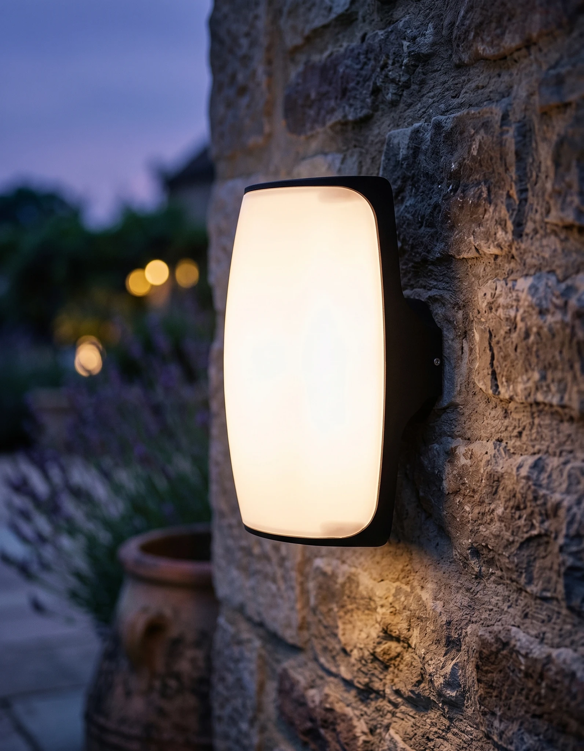 Outdoor wall sconce MAREA No.01 glowing on a rustic stone wall at dusk beside lavender and a terracotta pot