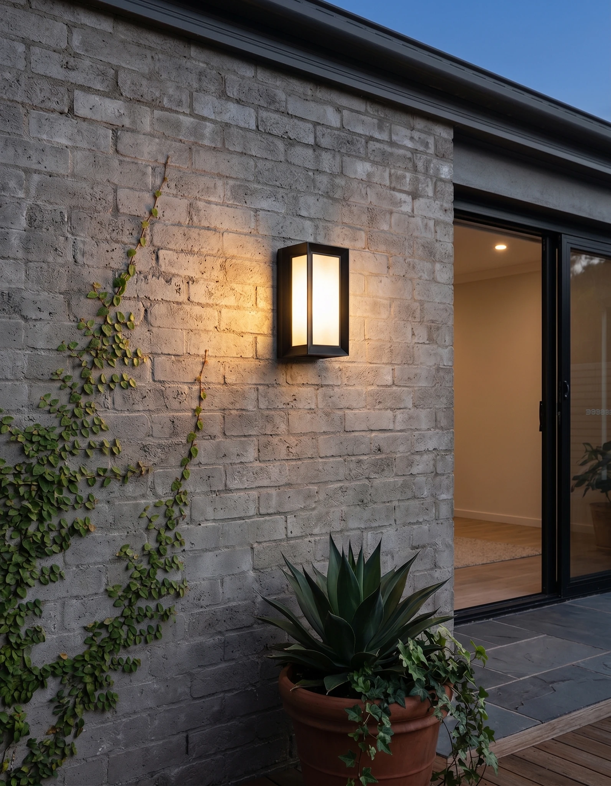 Outdoor wall sconce PORTICO No.01 lit on a white brick wall beside a sliding door with ivy and an agave planter