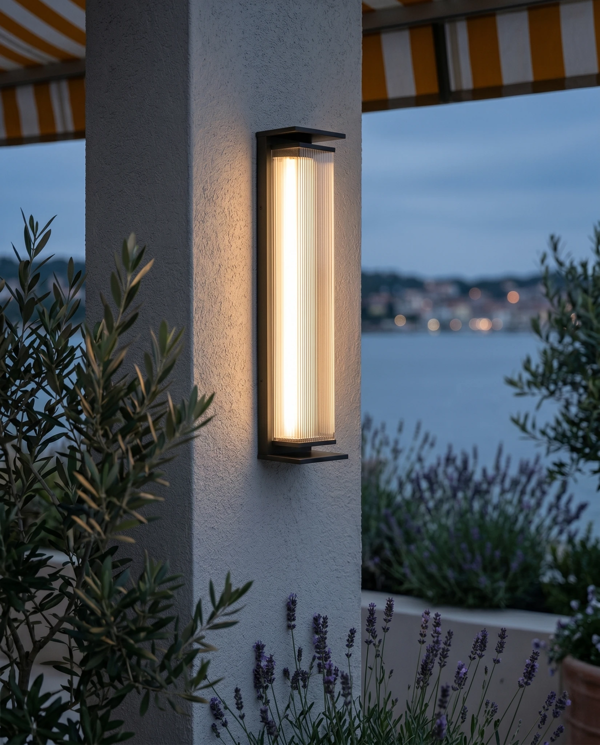 Outdoor wall sconce COSTA No.01 lit on a terrace wall at dusk with an olive tree, lavender and a sea view