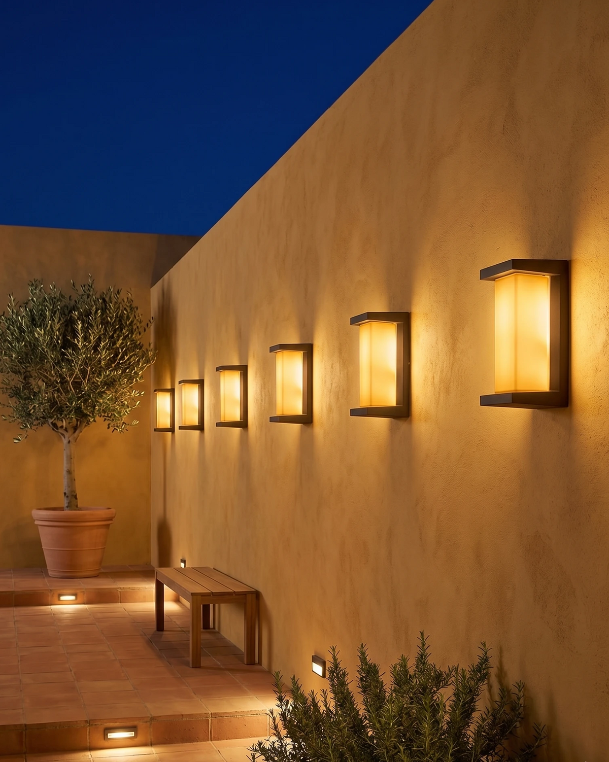 Five outdoor wall sconces PORTICO No.02 lit along an ochre stucco courtyard wall with an olive tree and bench