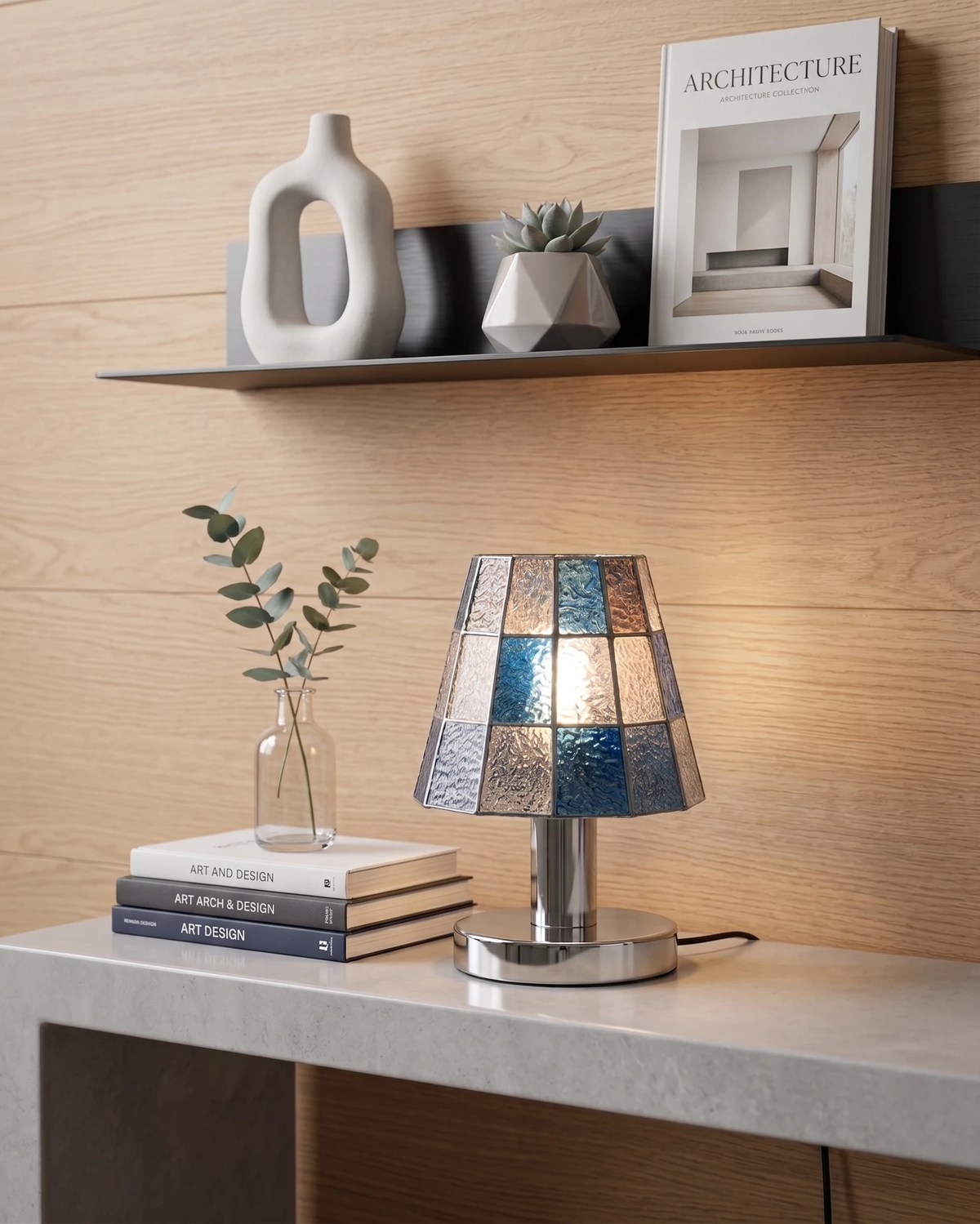 Stained glass table lamp VETRA No.01 in cobalt blue, lit on a white console below a shelf with books and a vase